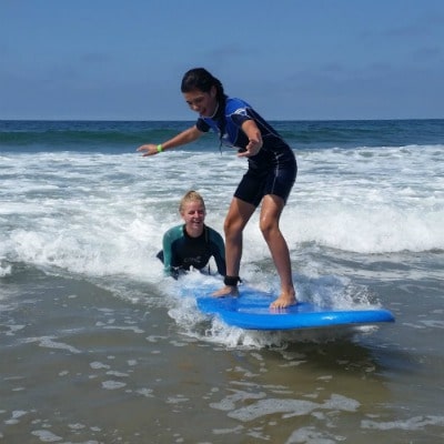 Camp counselor providing surfing instruction in the ocean to a teenage Aloha Beach Camp female camper.