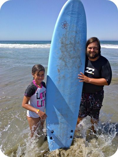 Surf camp director Matt Duda and a little girl he just taught how to surf standing in the ocean together holding a surfboard vertically between them.