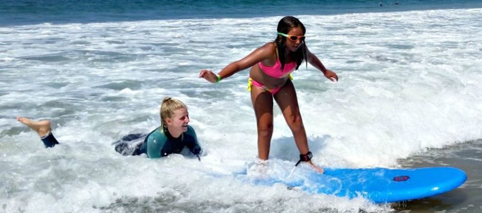 11 year old girl learning to surf at Aloha Beach Camp's Billabong Surf Camp at Zuma Beach in Malibu while her female surfing instructor holds the back of her board in the ocean to provide extra guidance, balance and support.