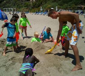 Camp counselor digging in the sand on the beach with several young campers in his group. All the kids have toy shovels and beach toys to play with.