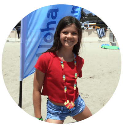 Young girl standing on the beach in front of an Aloha Beach Camp flag planted in the sand and wearing a candy lei necklace she made at Aloha Beach Camp.