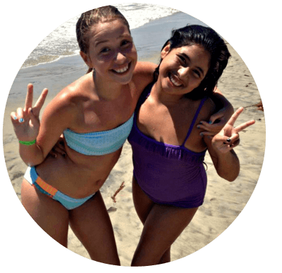 Two female Aloha Beach Camp campers standing on the beach at ocean's edge and giving 