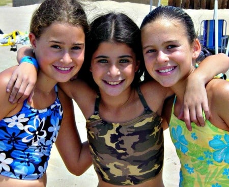 Three girls with arms around each other smiling and standing on the sand at Aloha Beach Camp.
