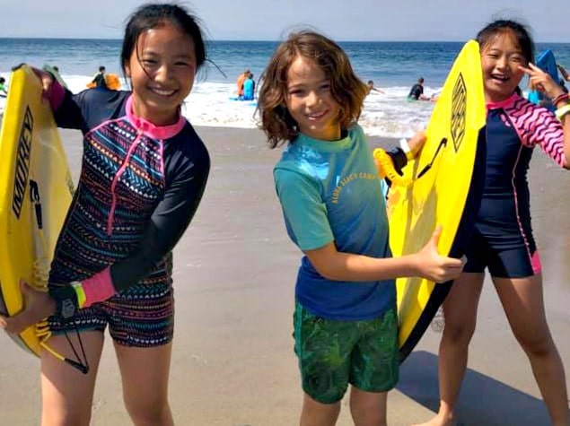 Three kids at summer camp together standing together on the wet sand holding boogie boards and smiling at Aloha Beach Camp in Los Angeles