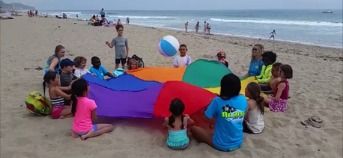 Campers and staff sitting in a circle on the sand playing with a colorful parachute and beach ball at Aloha Beach Camp Summer Camp.