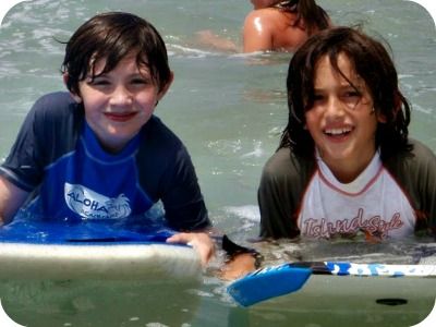 Two boys wearing Aloha Beach Camp rash guards laying side-by-side on their boogie boards in the ocean at Aloha Beach Camp.