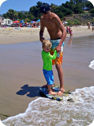Camp counselor standing on a boogie board with young camper on the shore, teaching him how to surf on the sand.