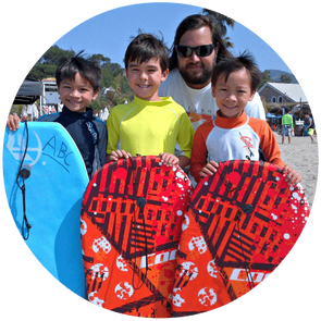 Three boys holding boogie boards standing on the beach with Aloha Beach Camp day camp director Matt Duda.