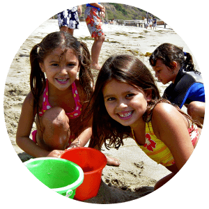 Three campers smiling, playing with their beach toys, and digging holes together in the sand at Aloha Beach Camp, L.A.'s only accredited summer beach day camp..