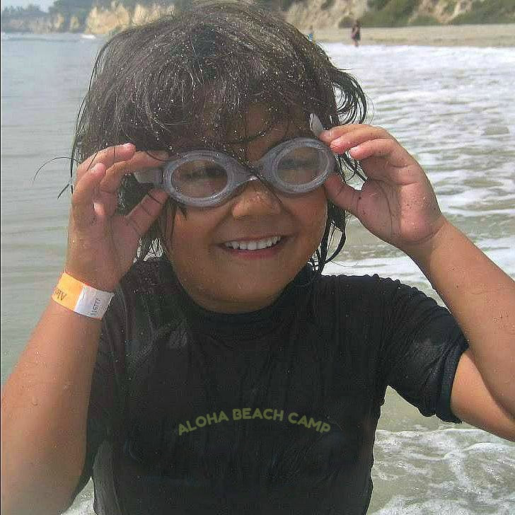 Boy wearing an Aloha Beach Camp rash guard standing in the Ocean at Paradise Cove adjusting his swimming goggles and smiling.