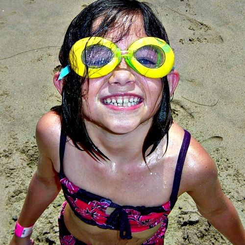 Young Keiki Camper girl wearing a pink bathing suit and bright yellow goggles standing on the sand at Aloha Beach Camp.