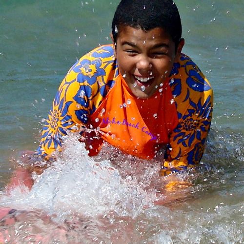 Happy smiling boy in bright orange rash guard with Aloha Beach Camp logo on the front boogie boarding at Aloha Beach Camp