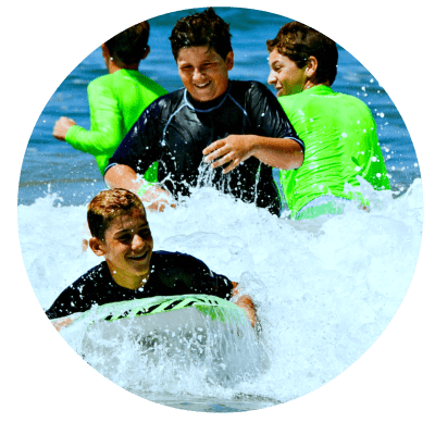 Teenage boys boogie boarding and laughing in the ocean at Aloha Beach Camp