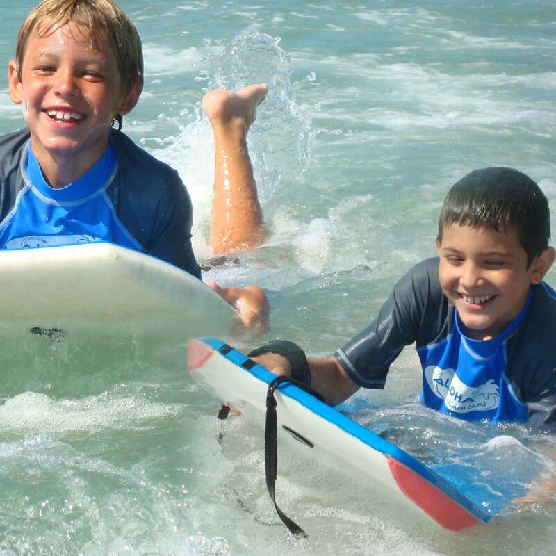 Two boys boogie boarding side-by-side together and smiling at Aloha Beach Camp's Kahuna Camp program at Zuma Beach, Malibu