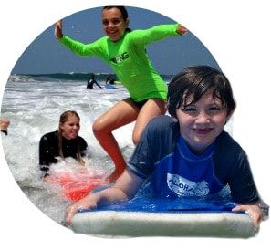 Boy boogie boarding wearing his Aloha Beach Camp rashgaurd while another camper surfs in the background with the help of her camp counselor surfing instructor.