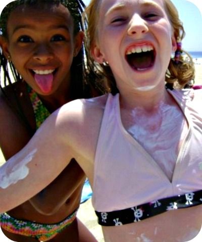 Two female campers laughing on the beach with their bodies covered in sunscreen.