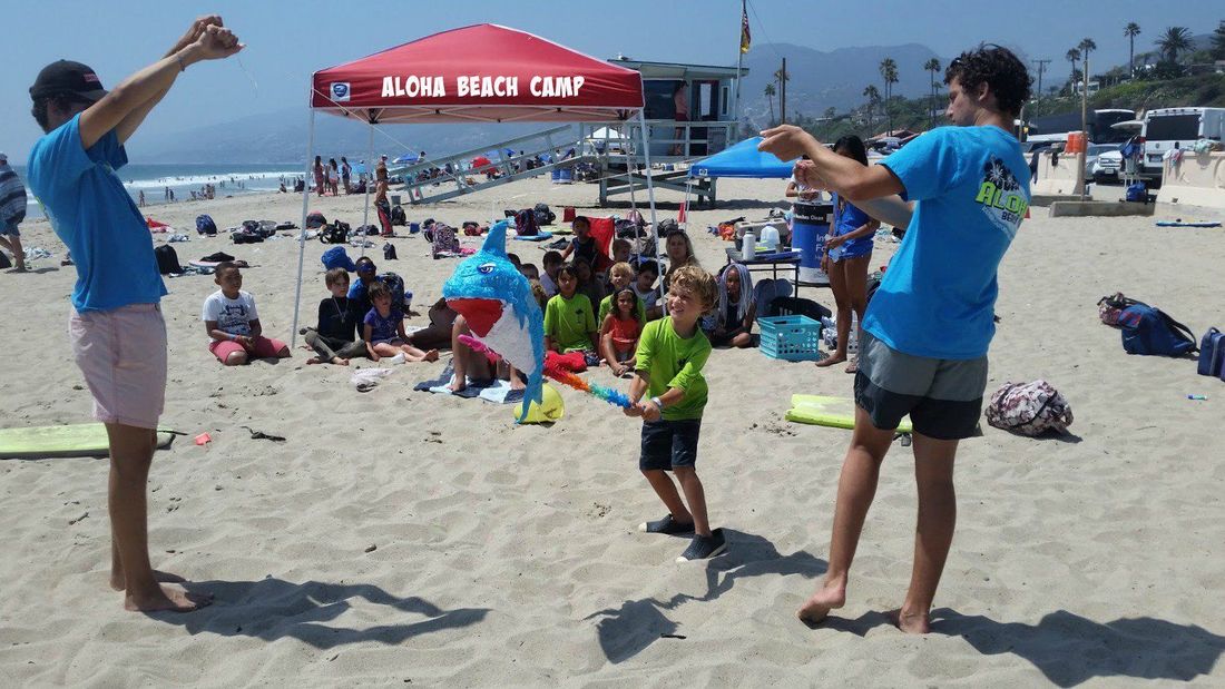 Two camp counselors holding a candy-filled shark pinata on the beach while a boy smacks the pinata with his stick as other campers look on.