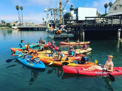 Campers and camp couselors sititing in their ocean kayaks holding paddles and smiling for the camera. A very colorful picture of at least 15 campers kayaking!
