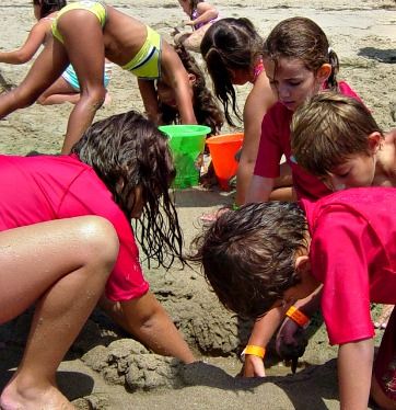 Group of campers in red rash guards digging in sand with beach toys at Aloha Beach Camp Summer Day Camp in Los Angeles.