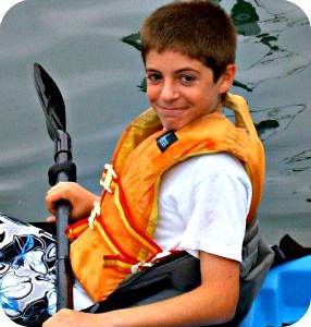 12 year old boy sitting in a blue kayak wearing an orange lifevest and holding his kayak paddle getting ready to enjoy Aloha Beach Camp's ocean kayaking summer camp activity with his friends. 