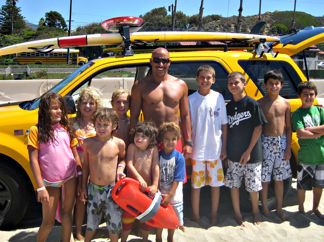 Zuma Beach Lifeguard with Aloha Beach Camp kids standing in front of lifeguard truck on the sand at Zuma Beach.