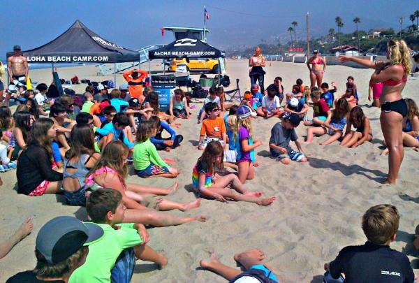 Camp directors, counselors and kids gathered together for a morning safety and assembly meeting on the beach at Aloha Beach Camp. About 40 campers are sitting on the beach while the staff stands and reviews the safety rules for the camp day.