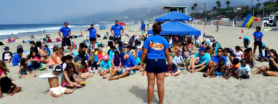Camp director Alxy Metcalf and other Aloha Beach Camp staff holidng an all-camp assembly safety meeting as campers sit on the beach and look on.