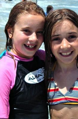 Two female campers standing together in the ocean and smiling. One is wearing a bathing suit, the other a pink and black Aloha Beach Camp rash guard. 