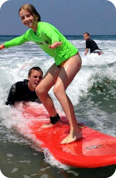 Teenage girl smiling and wearing a green rash guard while surfing on a red surfboard in the ocean at Aloha Beach Camp's Los Angeles surfing camp for kids and teens at Zuma Beach, while her surf camp instructor Shane McEvoy holds the back of the board to help her balance and guide her through the waves.