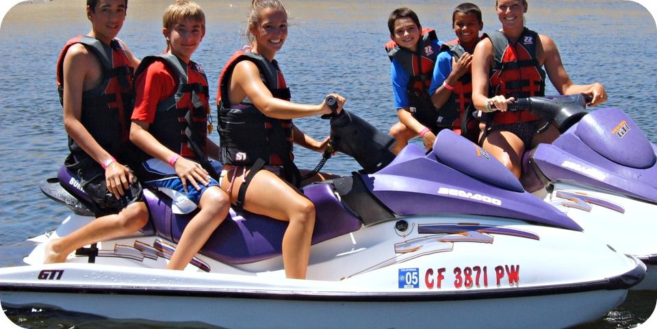 Two camp counselors, each driving a jet ski with two campers on the back of the jet skis, posing for a group photo at Aloha Beach Camp. 