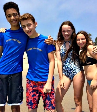 Four High Action Campers (two boys, two girls) standing together at the water's edge on the wet sand at Zuma Beach, Malibu while enjoying a day at Aloha Beach Camp's summer program.